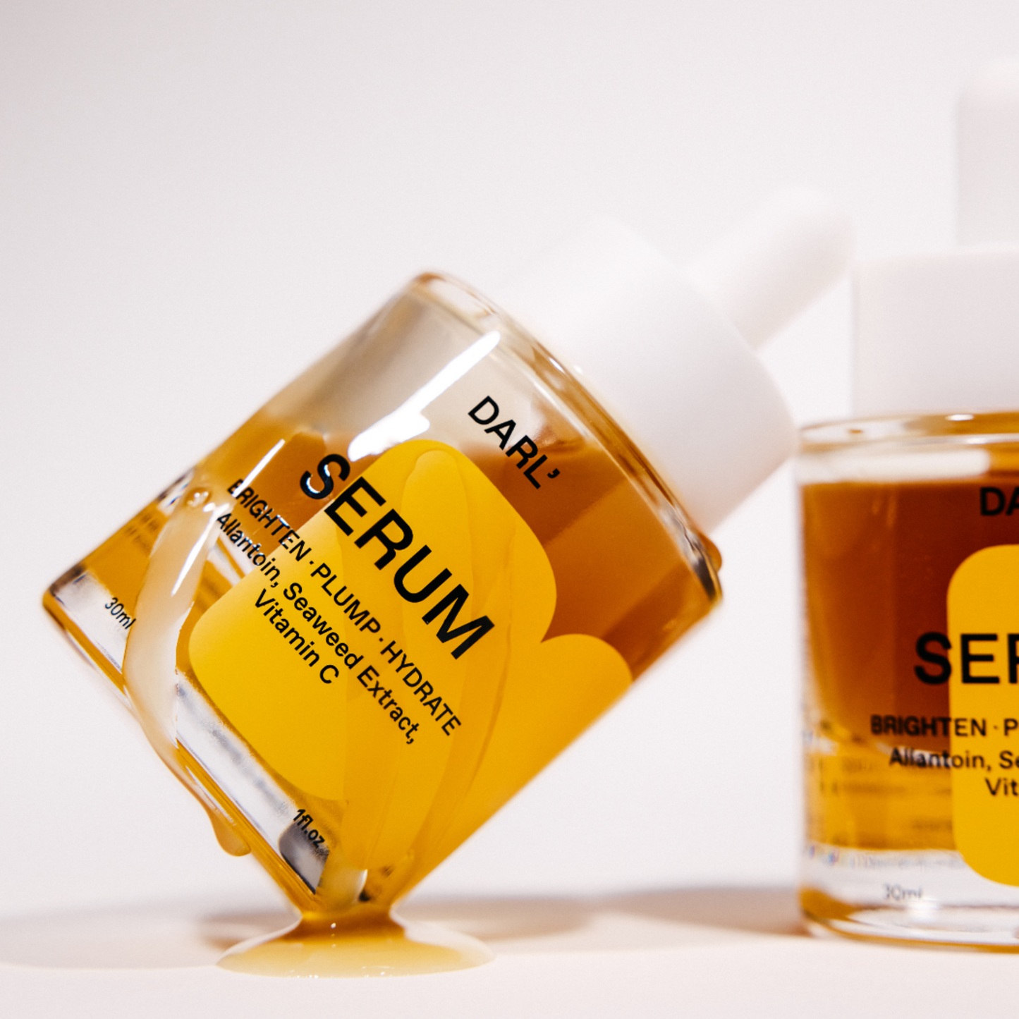 DARL' Brighten Plump Hydrate serum in a clear glass dropper bottle with a white cap, shown tipped over with amber liquid dripping onto a white surface. A second bottle is partially visible in the background.