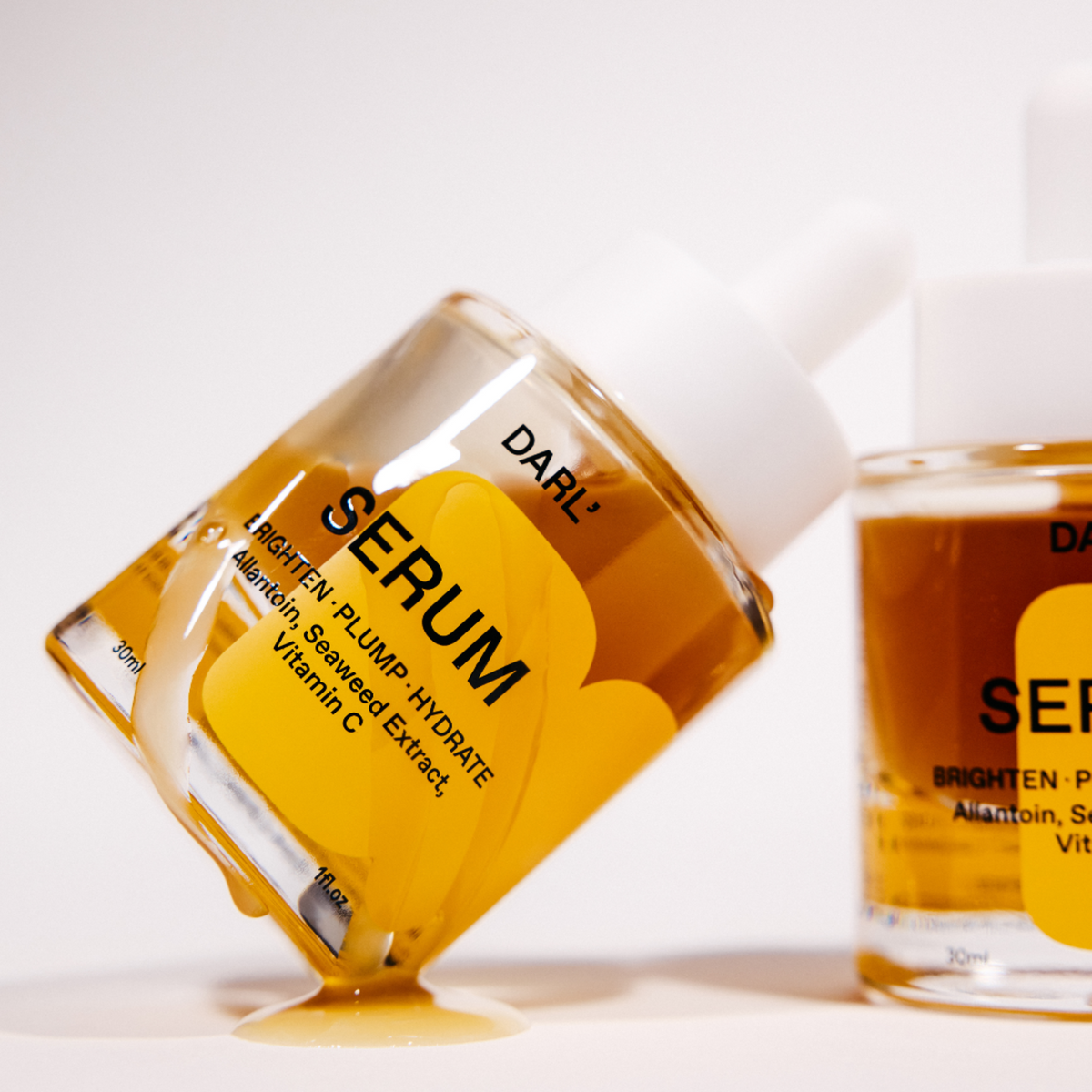 DARL' Brighten Plump Hydrate serum in a clear glass dropper bottle with a white cap, shown tipped over with amber liquid dripping onto a white surface. A second bottle is partially visible in the background.
