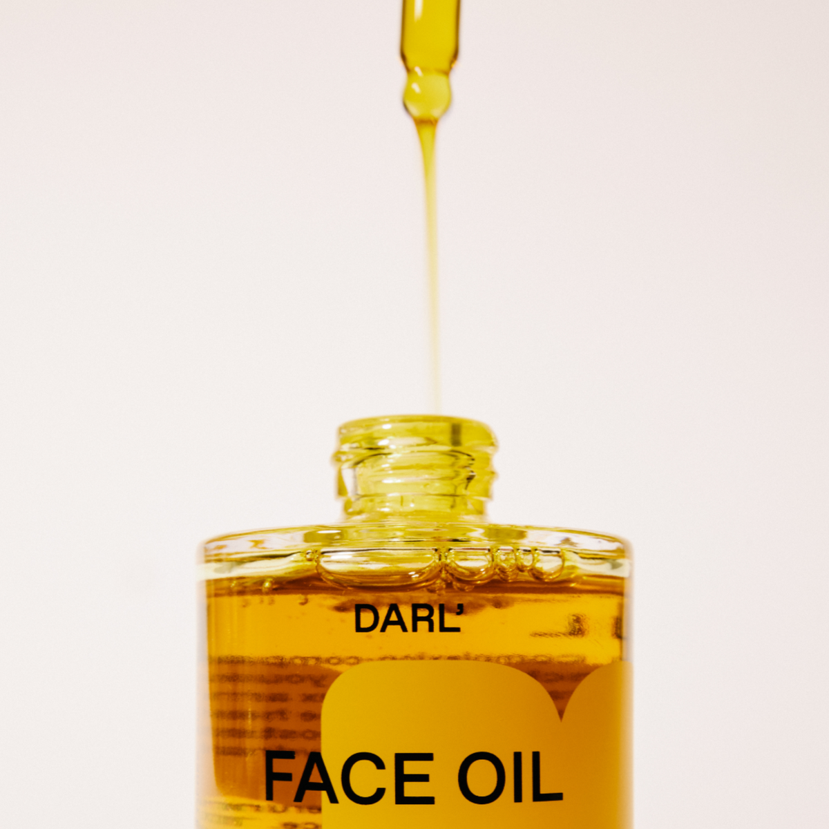 A close-up of a glass bottle of DARL' Face Oil with golden-yellow oil inside, showing a dropper above the open bottle with oil dripping into it against a clean white background.