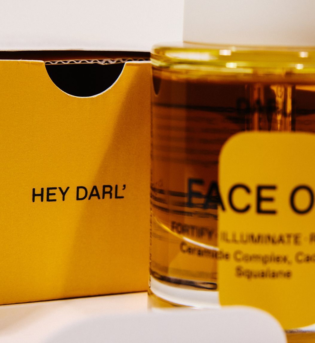 A close-up of a clear glass bottle filled with amber Darl' Face Oil liquid, next to a bright yellow box printed with 'HEY DARL', and a yellow label partially covering the bottle.